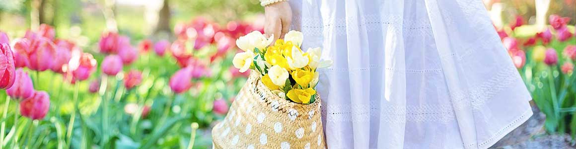 Person in weißem Sommerkleid hält geflochtene Tasche mit gelben Blumen, steht am Rand eines sonnigen Frühlingsfeldes voller rosa Tulpen