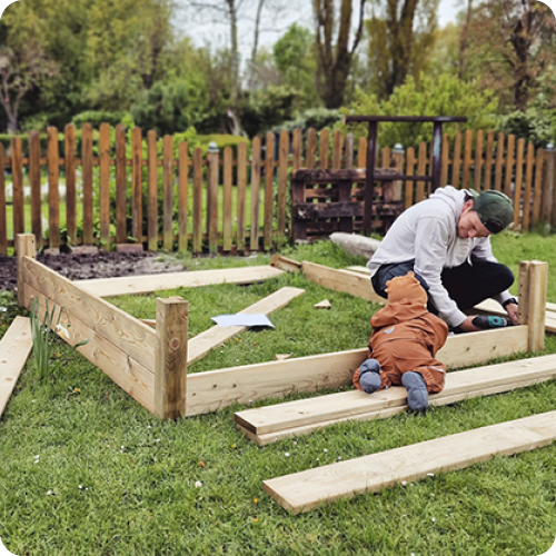 Erwachsener und Kleinkind bauen auf einer Rasenfläche einen Holzrahmen für ein Hochbeet, verstreute Holzbretter und Werkzeug, im Hintergrund ein Holzzaun und Bäume