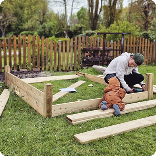 Erwachsener und Kleinkind bauen auf einer Rasenfläche einen Holzrahmen für ein Hochbeet, verstreute Holzbretter und Werkzeug, im Hintergrund ein Holzzaun und Bäume