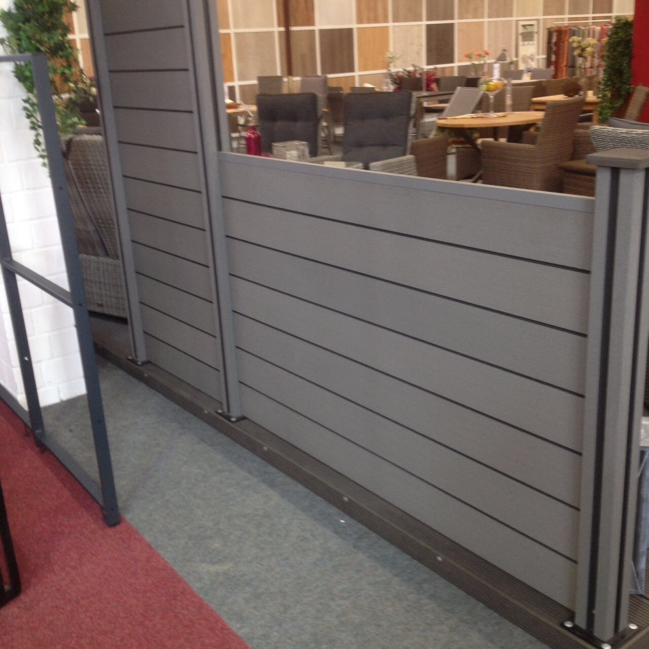 BPC Zaun-Stecksystem in Anthrazit, moderne Sichtschutzlösung für Garten oder Terrasse BPC Zaun-Stecksystem in Anthrazit, bestehend aus waagerecht verlaufenden Paneelen mit Pfosten, aufgestellt in einer Indoor-Ausstellung mit Gartenmöbeln im Hintergrund
