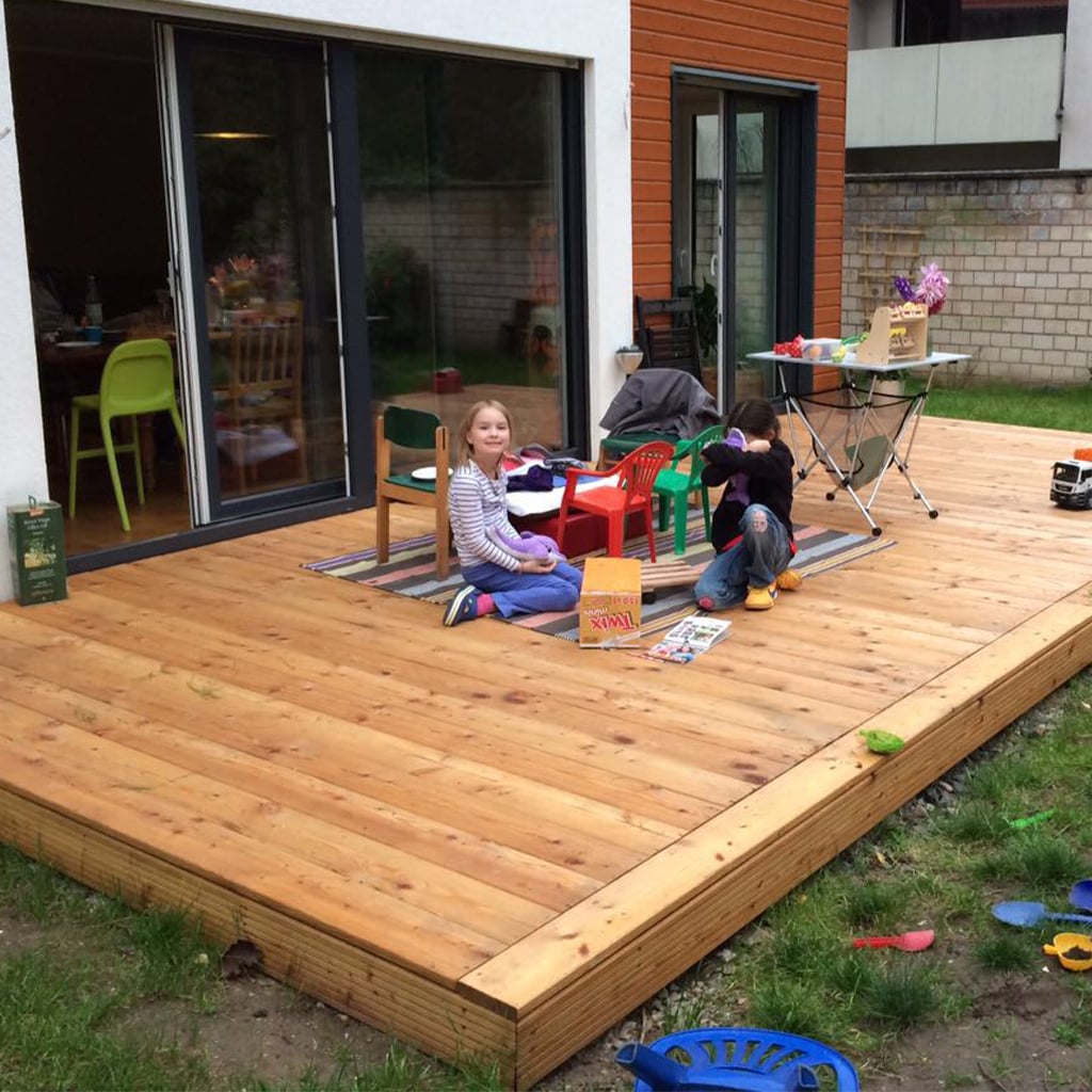 Zwei Kinder sitzen auf einer Holzterrasse vor einem modernen Haus mit großen Schiebetüren; bunte Kindermöbel, Spielzeug auf einem Teppich, ein Klapptisch mit Bastelmaterialien und verstreute Spielsachen im angrenzenden Gras