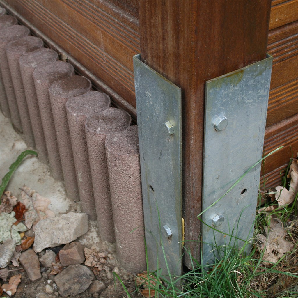 Winkelanker zum Einbetonieren 550 x 80 x 40 mm an Holzpfosten zur stabilen Befestigung im Außenbereich Winkelanker aus Metall zum Einbetonieren, befestigt an einem Holzpfosten, teils umgeben von Erde, Steinen, Gras und Betoneinfassung; Schrauben sichern den Anker am Holzpfosten