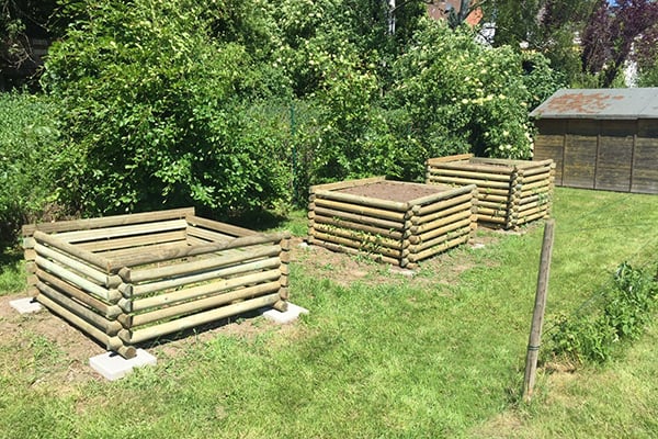 Drei quadratische Hochbeete aus übereinander geschichteten Rundhölzern auf Rasen, im Hintergrund Büsche und ein kleiner Holzschuppen bei sonnigem Wetter