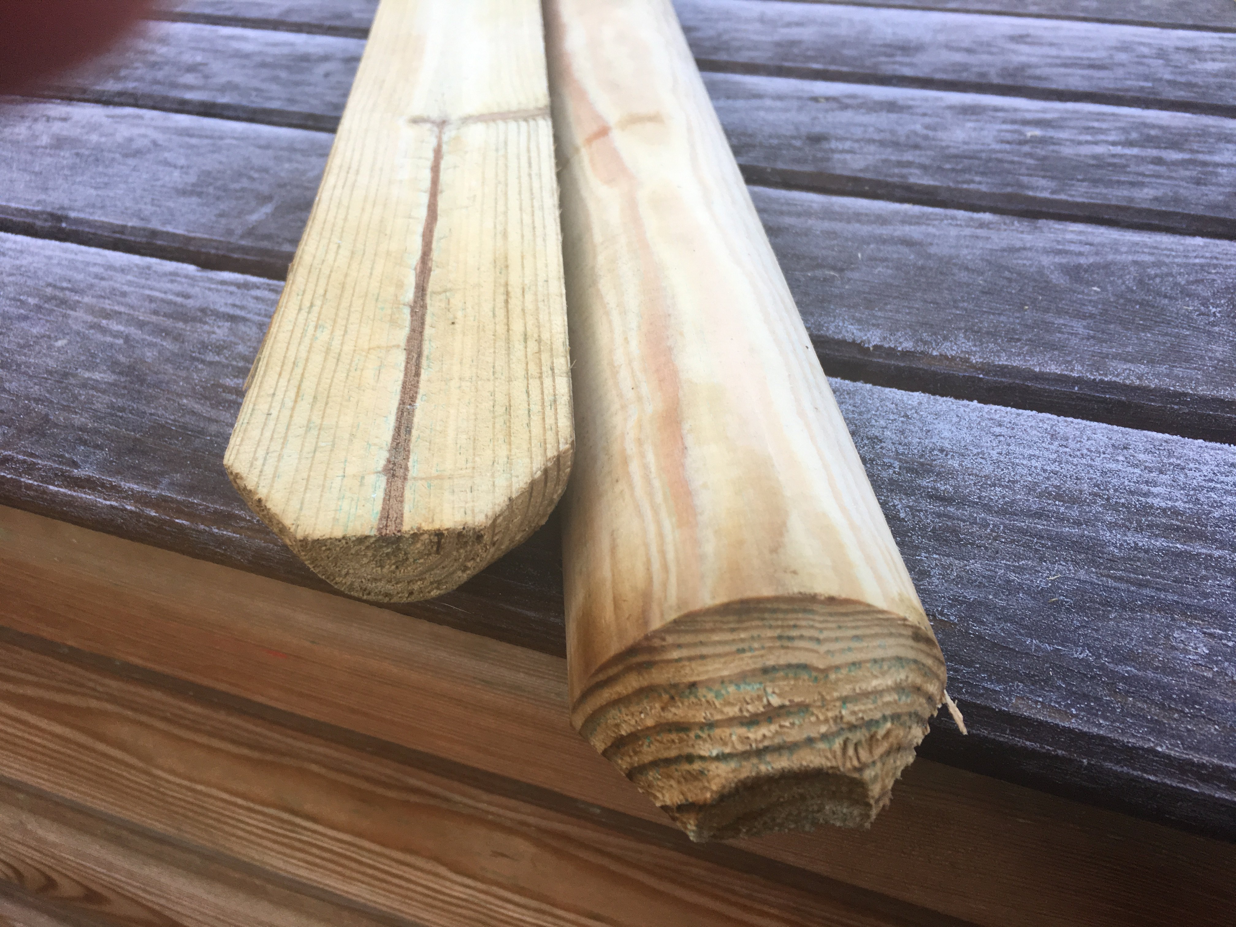 Zwei gekegelte, naturfarbene, kesseldruckimprägnierte Holzstaketen (6 cm stark) in Nahaufnahme, liegend auf wettergealterten Holzbrettern, beide Staketen mit deutlich sichtbarer Maserung und unterschiedlichen Zuschnittformen