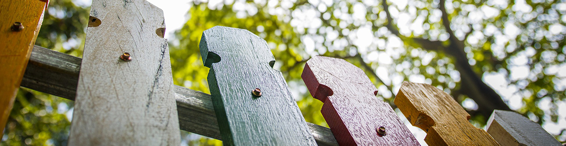Bunte, schräg fotografierte Holzlatten eines Zauns (weiß, grün, rot, gelb) mit sichtbaren Schrauben vor unscharfem Laub und Sonnenlicht