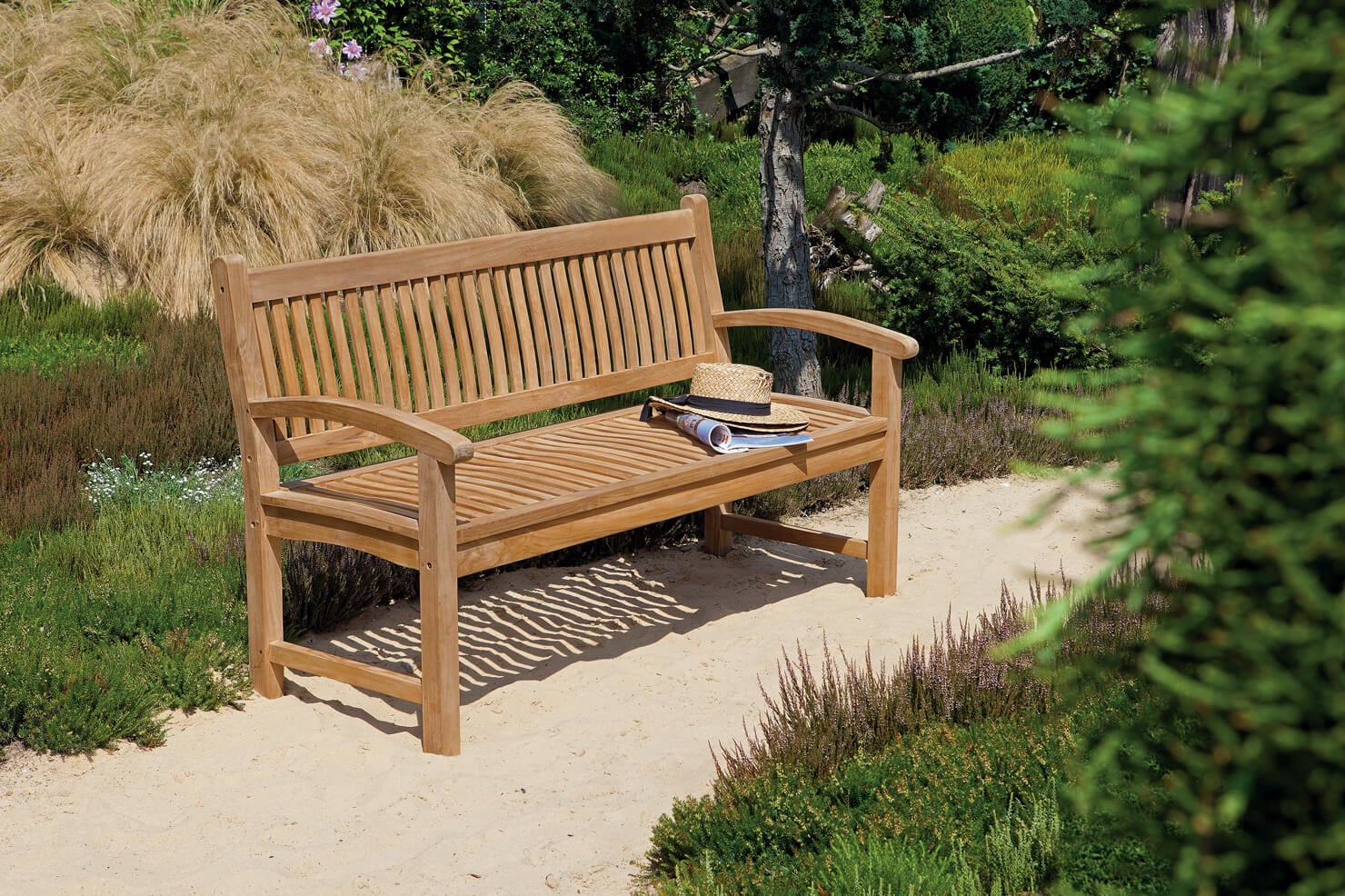 Garten Teakholzbank auf sandigem Weg in einem sonnigen Garten, auf der Sitzfläche ein Strohhut und ein aufgerolltes Magazin, umgeben von Gräsern, Heidekraut, Büschen und einem Baum, Schatten der Latten auf dem Sand