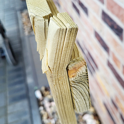 Nahaufnahme einer gesplitterten Holzlatte eines Zauns mit freigelegten Schichten und Splittern, unscharfer Gehweg und Backsteinwand im Hintergrund