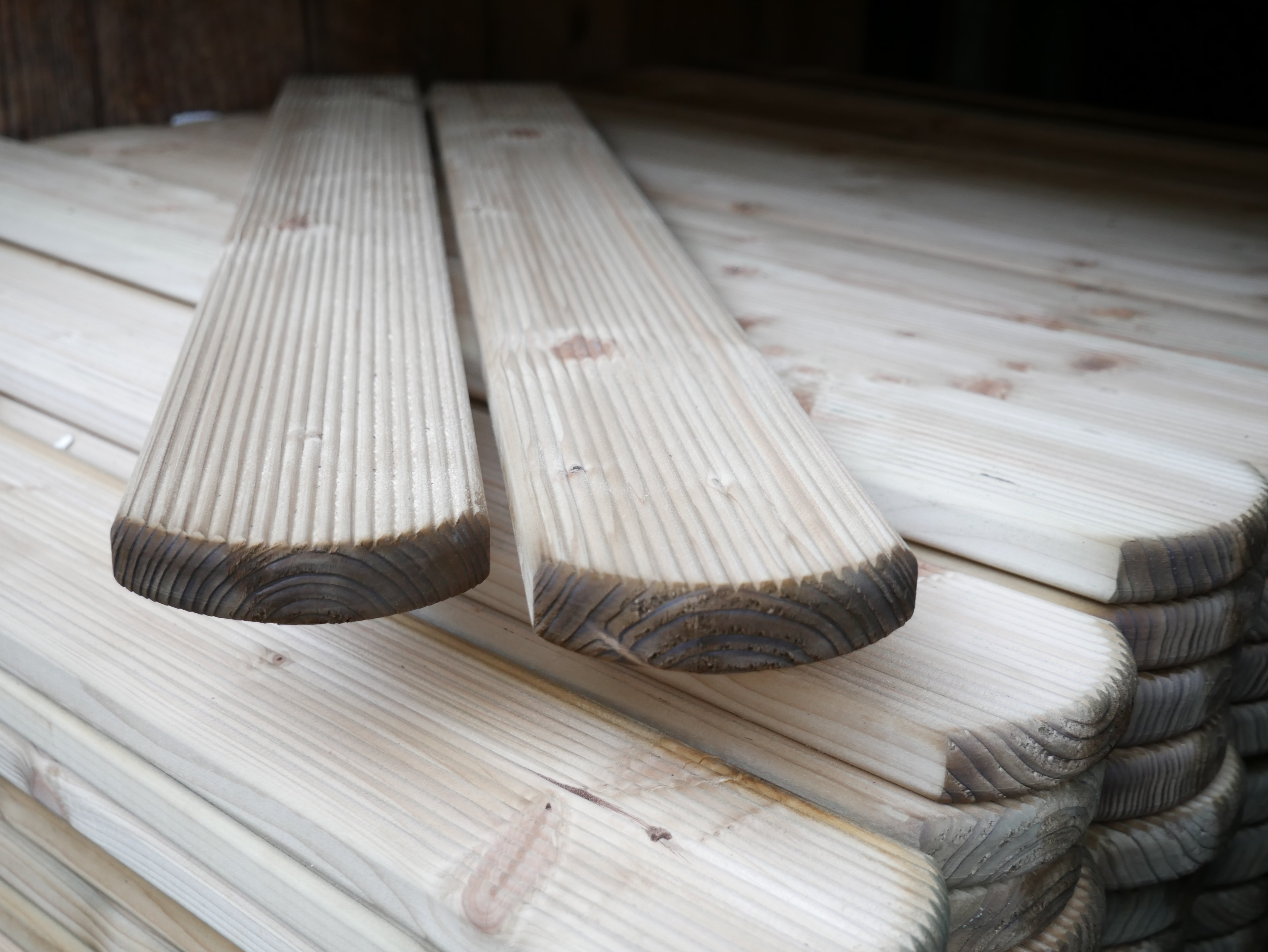 Mehrere kesseldruckimprägnierte, geriffelte Zaunbretter vom Typ Mosel mit abgerundetem Kopf, Format 22 x 95 mm, liegen gestapelt übereinander auf einer Holzfläche.