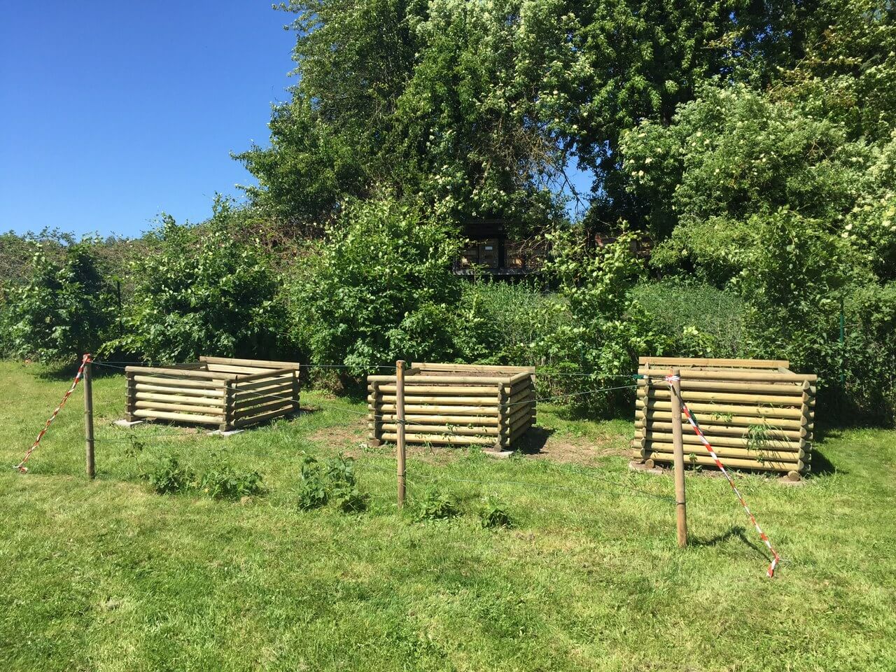 Drei rechteckige, aus Rundhölzern gebaute Kompostbehälter auf einer Wiese, dahinter dichter Strauch- und Baumbestand und blauer Himmel, im Vordergrund Pfähle mit Absperrband und Seil