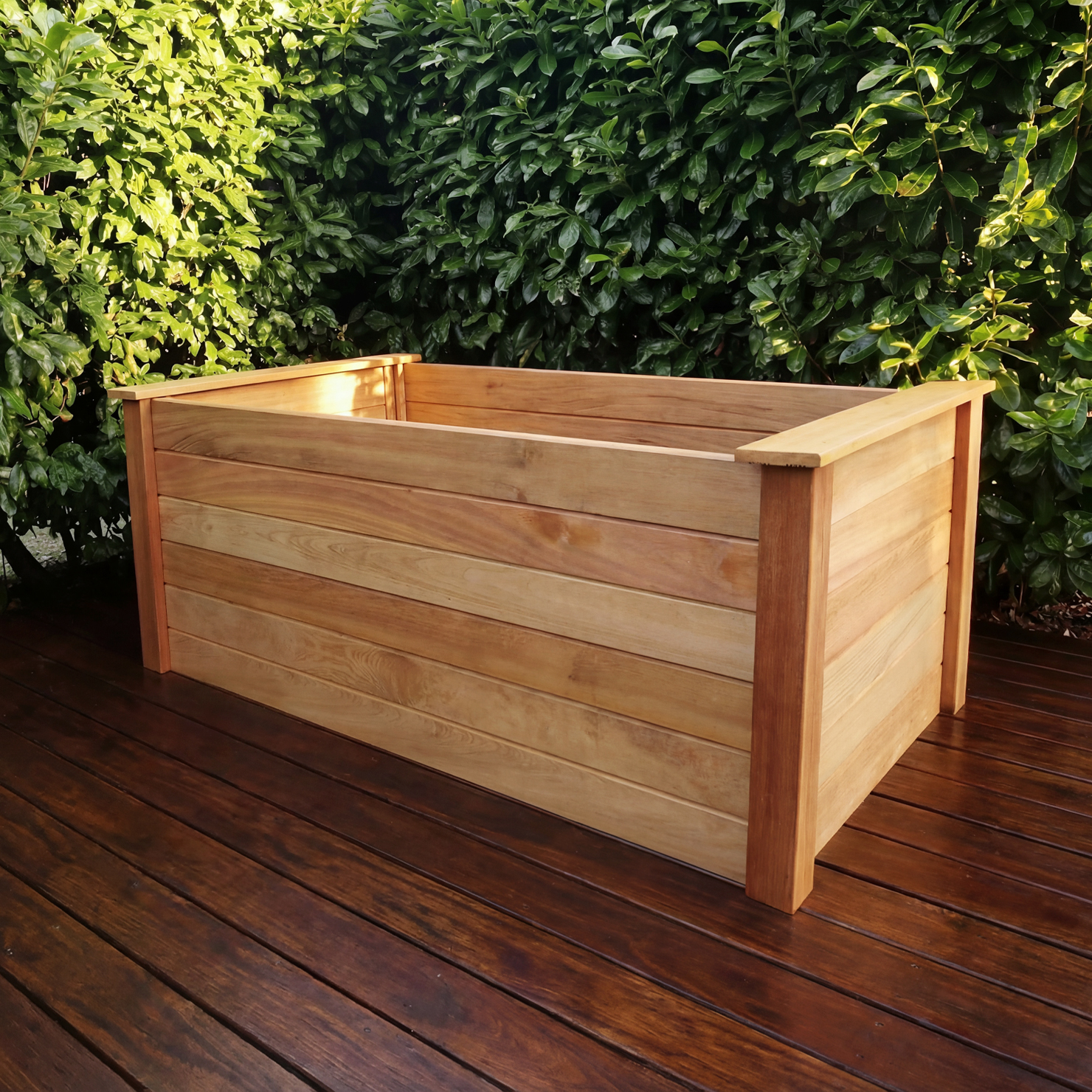 JULIA‑Hochbeet Murure kurze Seite, naturfarbenes rechteckiges Holzhochbeet mit horizontalen Leisten und breiten Pfosten auf dunklem Holzdeck vor grüner Hecke