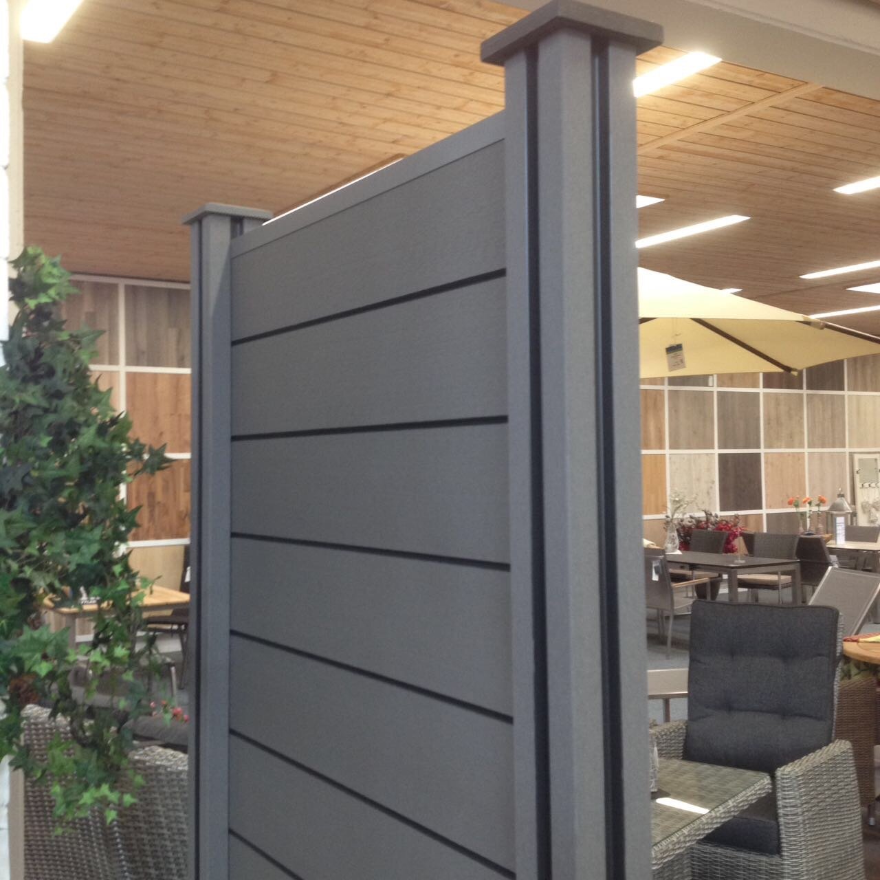 BPC Zaun-Stecksystem in der Farbe Anthrazit, montiert in einem Ausstellungsraum mit Holzwand und Gartenmöbeln im Hintergrund, moderne Optik mit waagerechten Paneelen und zwei Pfosten