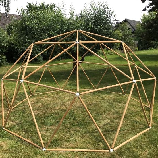 Freistehendes Holzgerüst einer geodätischen Kuppel aus dünnen Rundstäben auf Rasen, im Garten vor Bäumen und Wohnhäusern im Hintergrund