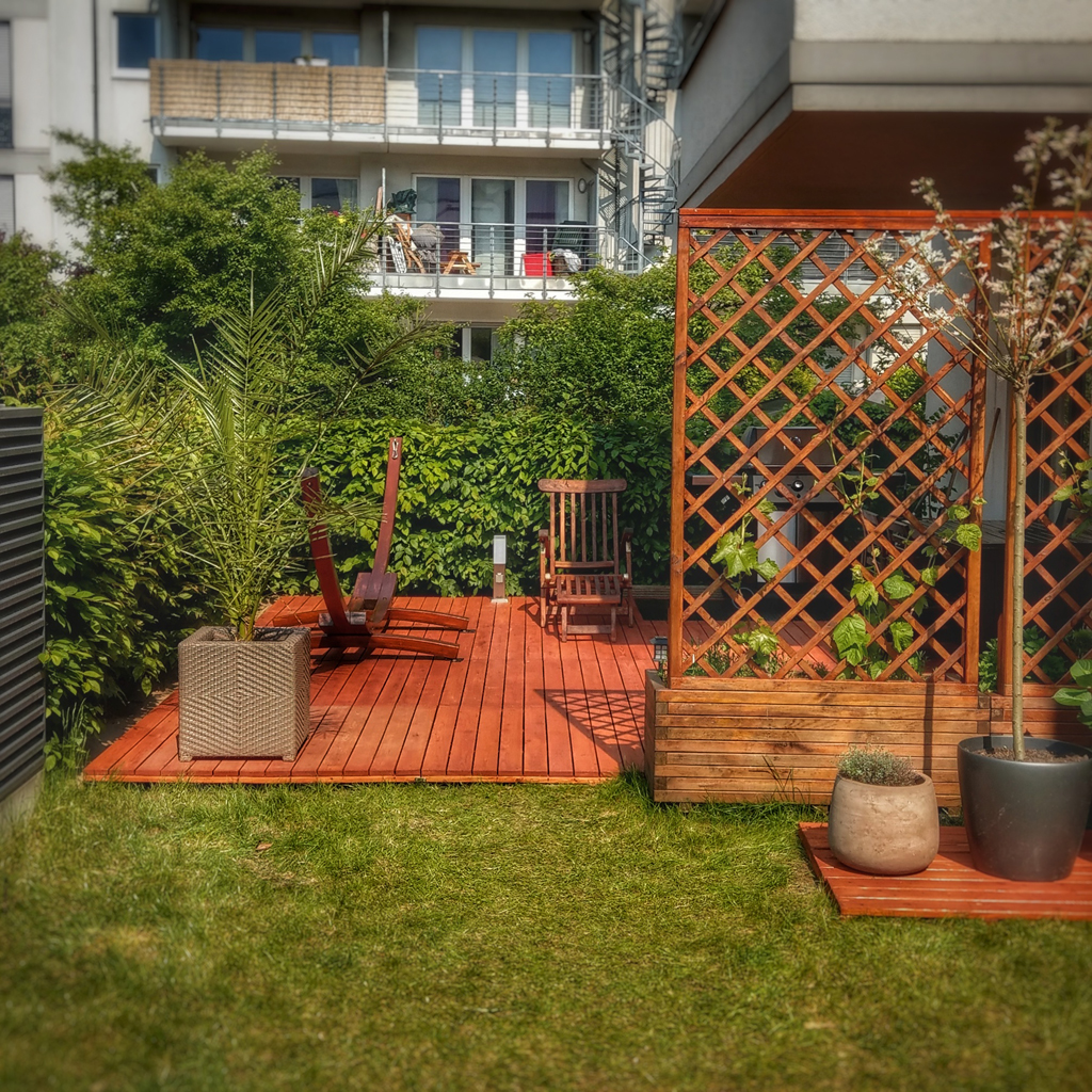 Kleiner Garten mit rotem Holzdeck, Rankgitter an der Seite, zwei Blumentöpfen, Palme in Korb, Liegestuhl und Holzstuhl auf der Terrasse, dichte Hecke im Hintergrund und Wohnhaus mit Balkonen darüber