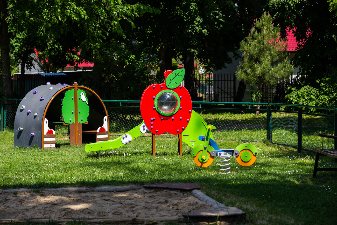 Kinderrutsche in Apfelform, 240 x 80 cm, auf einem Spielplatz. Bunte Kinderrutsche in Apfelform auf Rasen in einem Spielplatz, umgeben von Bäumen, mit einer Sandfläche im Vordergrund und einem Klettergerüst im Hintergrund.