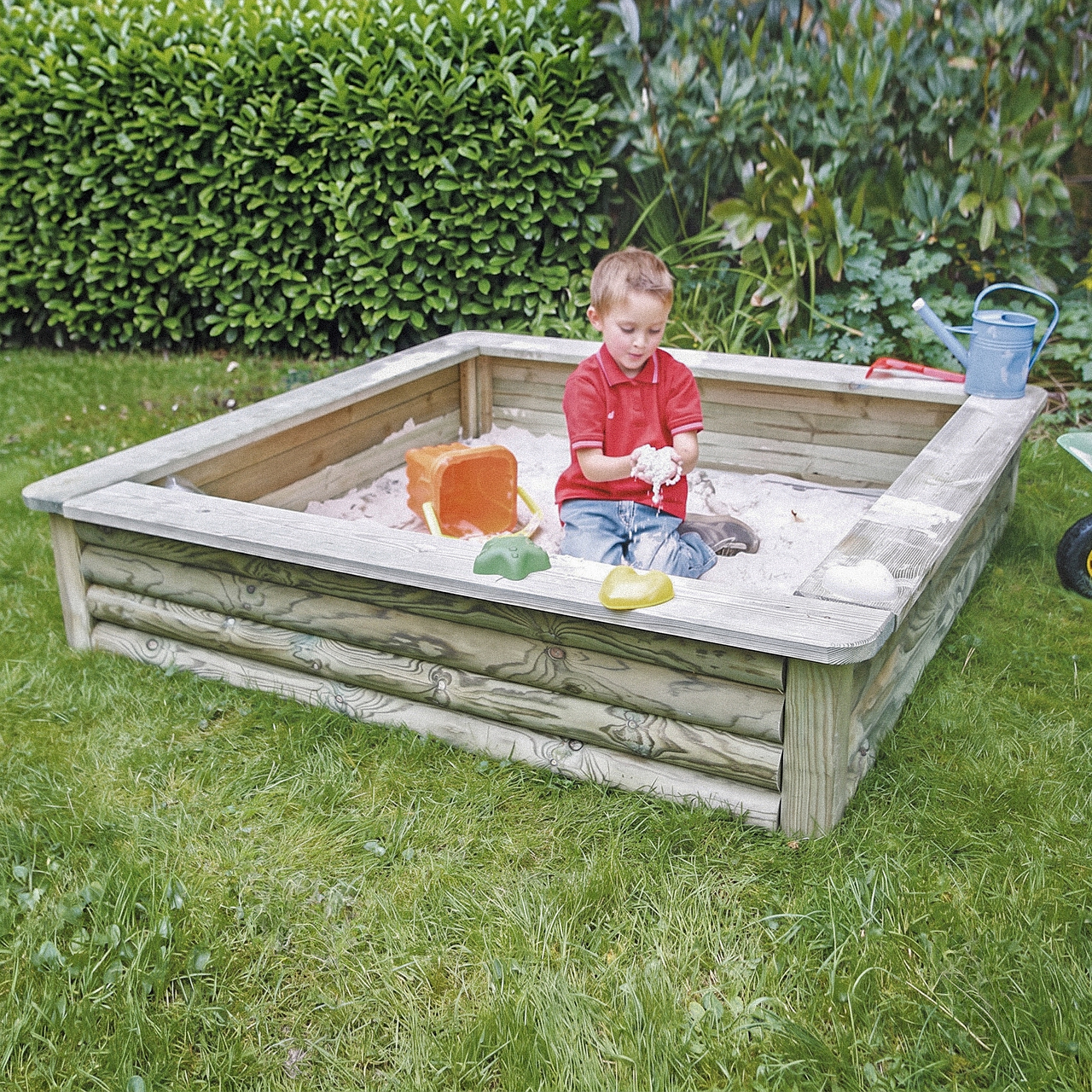 Ein Kind sitzt in einem hölzernen Sandkasten im Garten und spielt mit Sand, trägt rotes Polohemd und Jeans, umgeben von einem orangefarbenen Eimer, grünen und gelben Sandförmchen und einer blauen Gießkanne am Rand, Hecke im Hintergrund