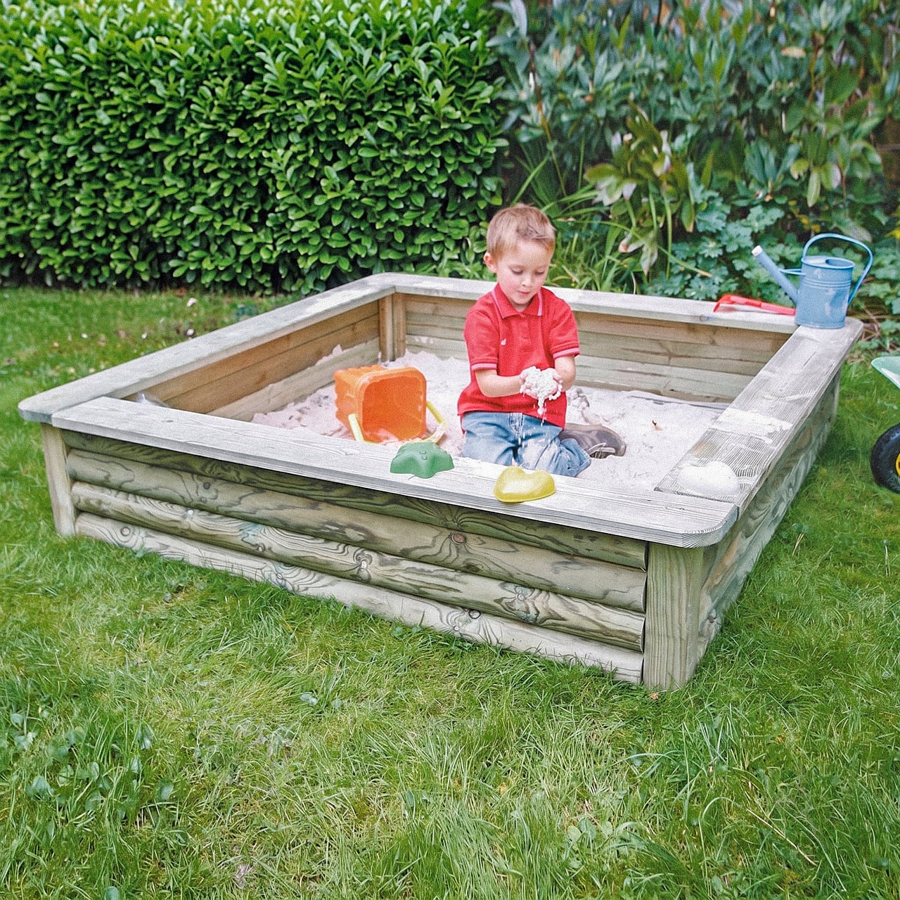 Sandkasten Tommy 150 x 150 x 27 cm Ein Kind sitzt in einem hölzernen Sandkasten im Garten und spielt mit Sand, trägt rotes Polohemd und Jeans, umgeben von einem orangefarbenen Eimer, grünen und gelben Sandförmchen und einer blauen Gießkanne am Rand, Hecke im Hintergrund