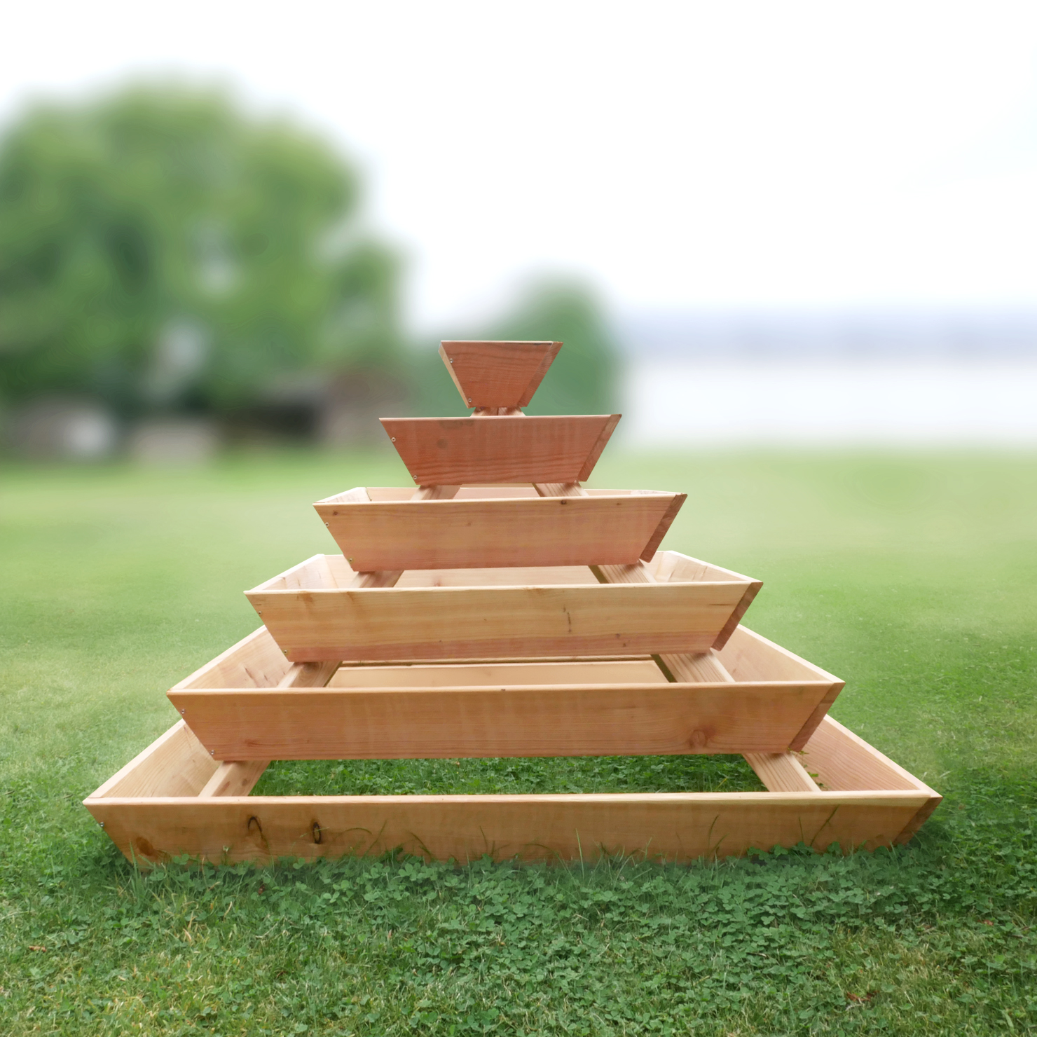 Holzpyramide aus Lärche, naturbelassen, bestehend aus fünf Ebenen, leer, auf einem Rasen. Geeignet für Kräuterbepflanzung. Maße: 120 x 120 x 87 cm. Hintergrund unscharf mit Bäumen und Himmel.