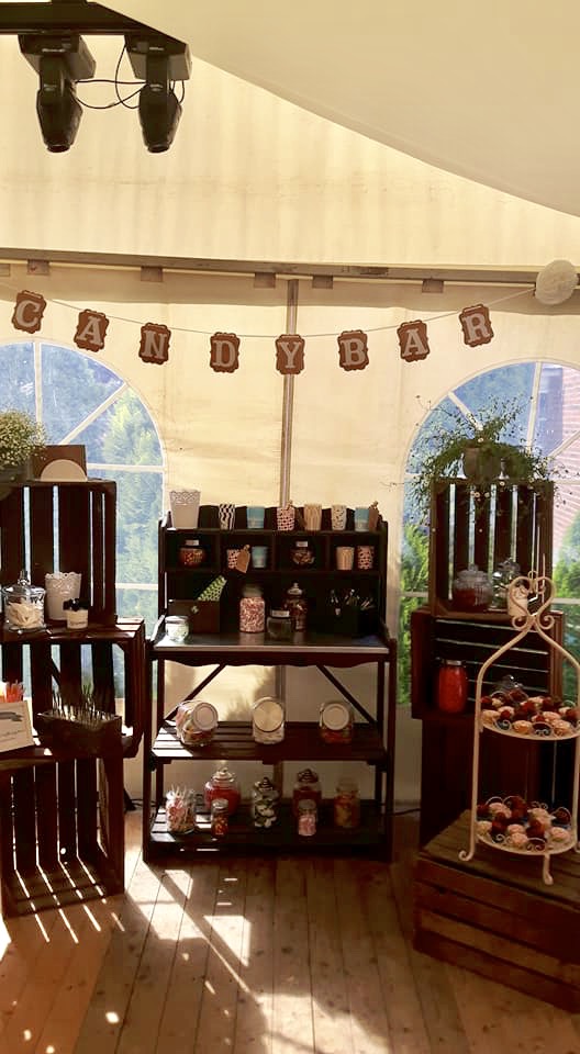 candybar Zelt-Candybar mit Girlande CANDYBAR, hölzernen Kisten und Regalen mit Bonbongläsern, Schalen und süßen Leckereien, zwei Bogenfenstern im Hintergrund, Holzboden und Bühnenbeleuchtung an der Decke
