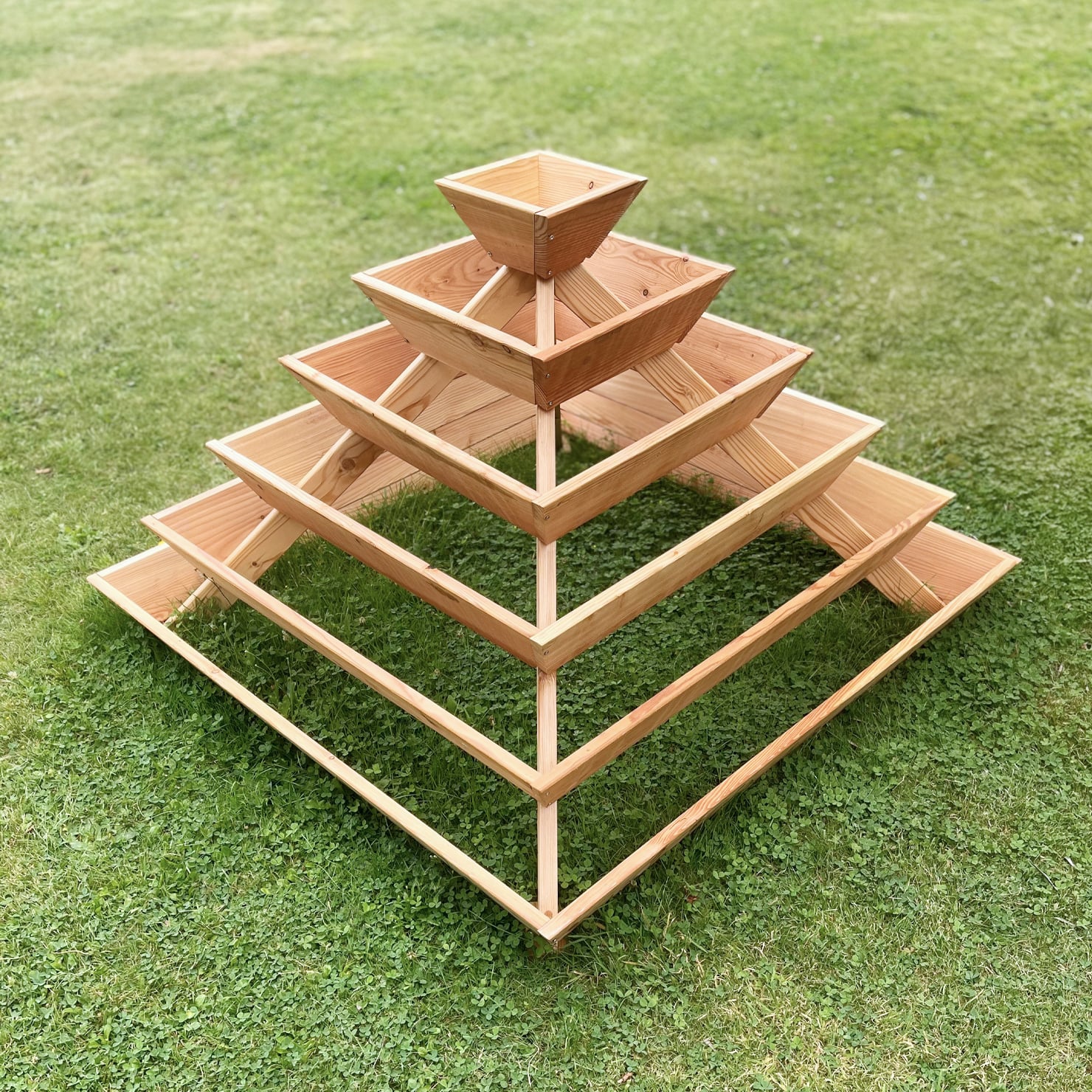 Kräuter-Pflanzenpyramide aus Lärchenholz auf Rasenfläche Pyramidenförmige Pflanzenstruktur aus naturbelassenem Lärchenholz mit mehreren Ebenen auf einer Rasenfläche, geeignet für Kräuteranbau, inklusive Vlies, Abmessungen 120 x 120 x 87 cm.