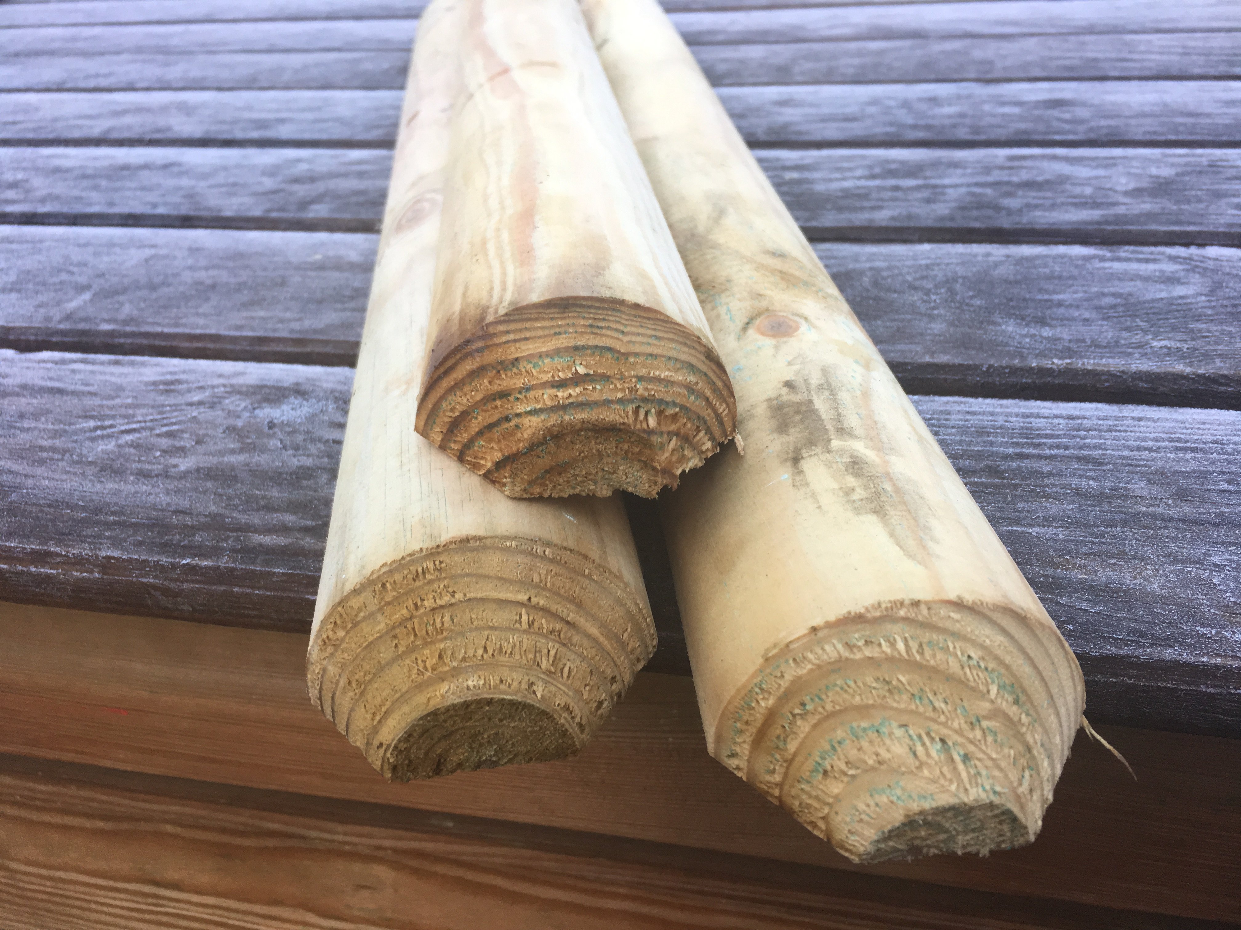 Gekegelte, kesseldruckimprägnierte Staketen aus Holz, 6 cm stark, in naturbelassener Ausführung Drei gekegelte, 6 cm starke, kesseldruckimprägnierte Holzstaketen in Naturfarbe, liegend auf einer Holzoberfläche, Nahaufnahme der Stirnseiten