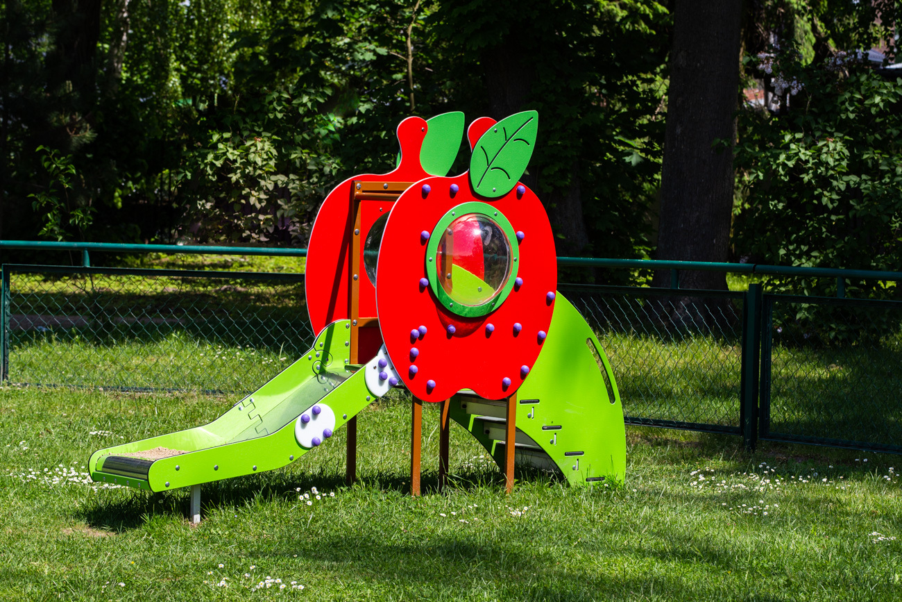 Kinderrutsche in Form eines Apfels, rot und grün, mit kleinem Kletterbereich, auf grünem Rasen im Freien. Maße: 240 x 80 cm.
