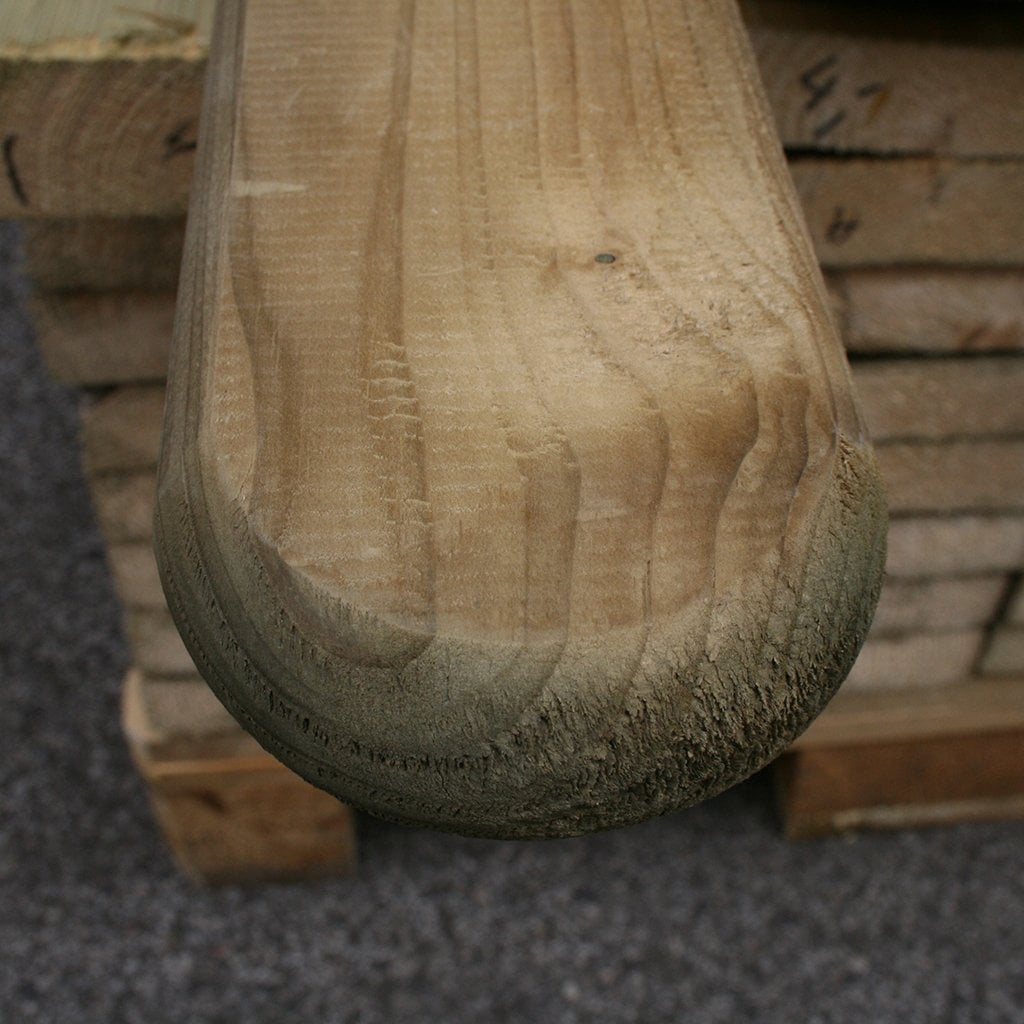 Zaunbrett Schonberg mit Rundkopf, stark gefast, kesseldruckimprägniert, 24 x 90 mm Nahaufnahme eines kesseldruckimprägnierten Zaunbretts Schonberg mit Rundkopf, stark gefasten Kanten, Maß 24 x 90 mm; im Hintergrund gestapelte Holzbretter, Untergrund aus Asphalt