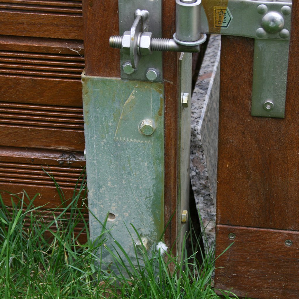 Detailansicht eines Winkelankers zum Einbetonieren (550 x 80 x 40 mm) aus Metall, montiert an einem Holztor, teilweise von Gras umgeben, mit sichtbaren Schrauben und Scharnieren zur Befestigung