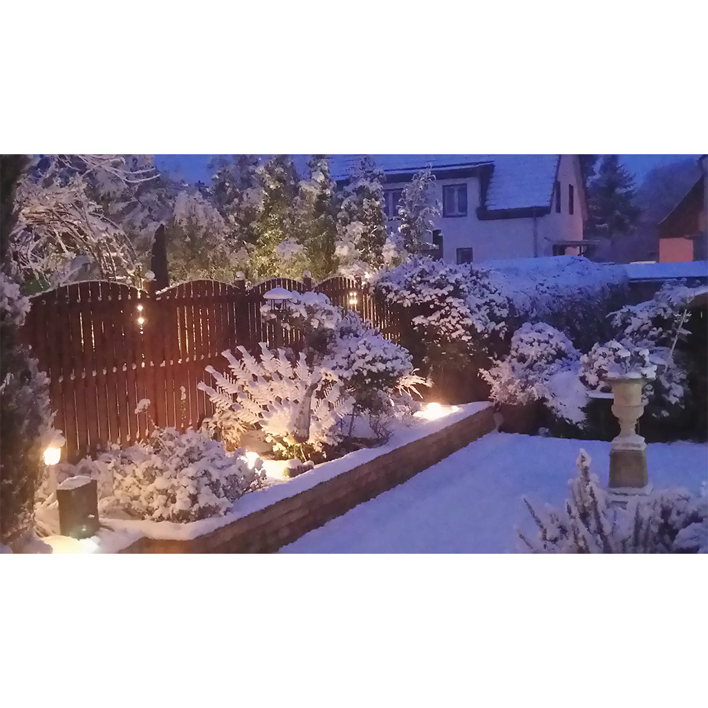 Verschneiter Hausgarten bei Dämmerung mit warmen Bodenspots, schneebedeckten Beeten und Sträuchern, einem dunklen Holzzaun, im Hintergrund ein Haus und rechts im Vordergrund eine steinerne Säule/Urne