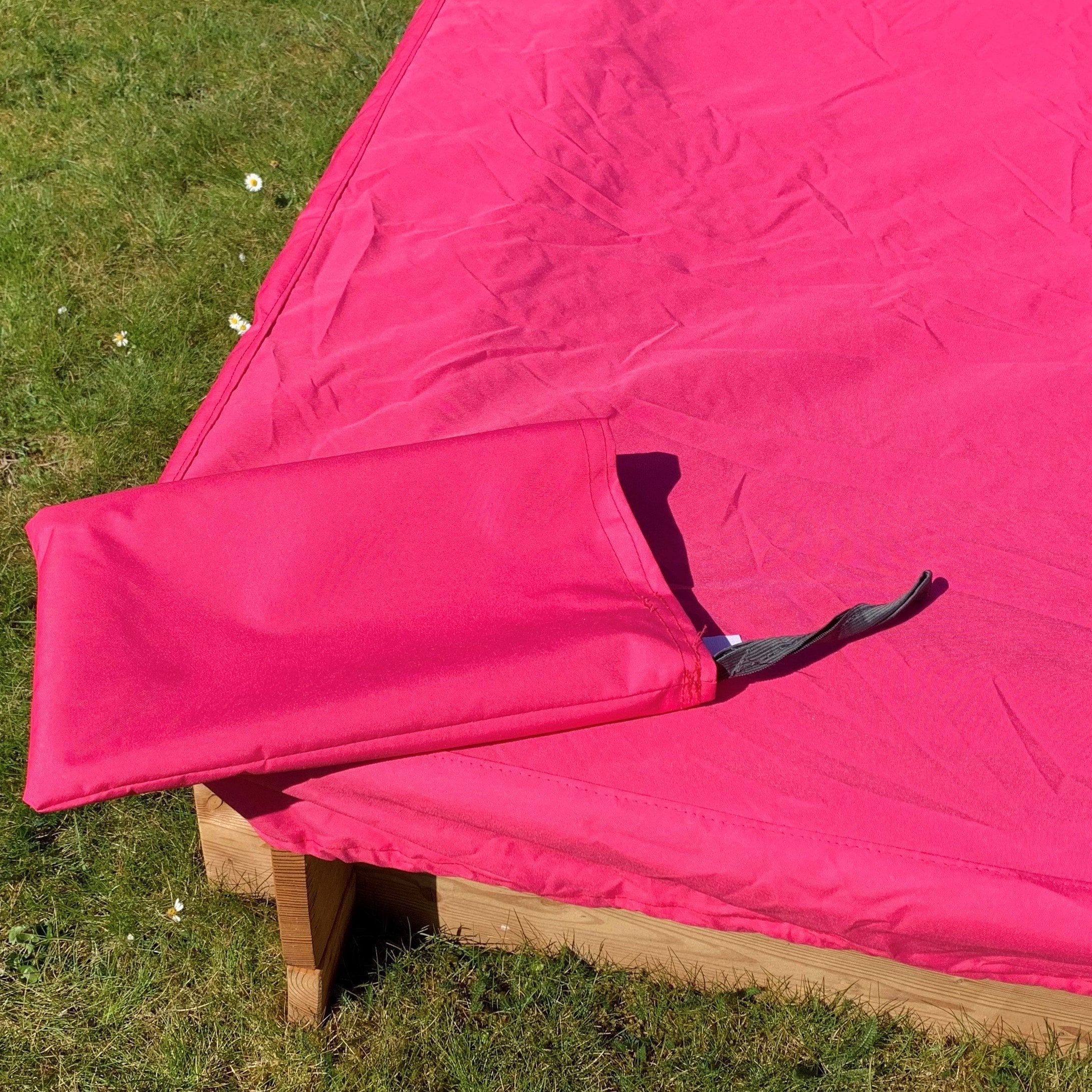 Rote Abdeckplane schützt Sandkasten vor Schmutz und Wetter Rote Abdeckplane auf einem Holzsandkasten im Garten, eine Ecke der Plane ist umgeschlagen und zeigt einen angenähten Befestigungsriemen, grüner Rasen mit Gänseblümchen im Hintergrund