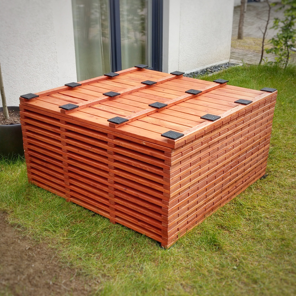 Rechteckiger Stapel aus roten Holzlatten auf Rasen vor einer Hauswand und Tür, Seiten mit gleichmäßigen horizontalen Schlitzen und oben mehrere kleine schwarze Gummipads