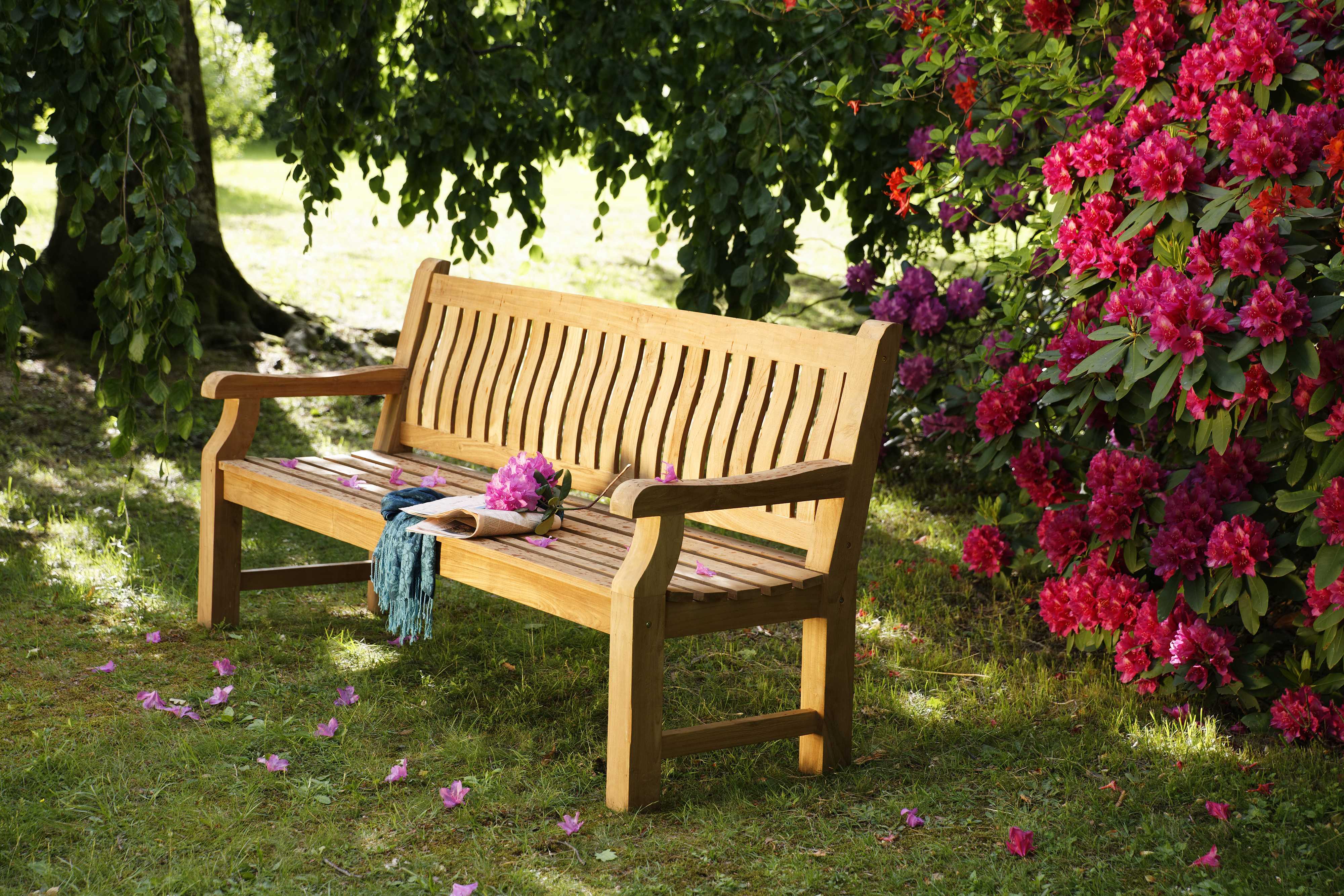 Holzbank aus recyceltem Teak, 2-3 Sitzer, im Garten auf Gras, flankiert von blühendem pinken Rhododendron und einem großen Baum im Hintergrund. Auf der Bank liegt ein rosa Blumenstrauß und ein türkisfarbener Schal.
