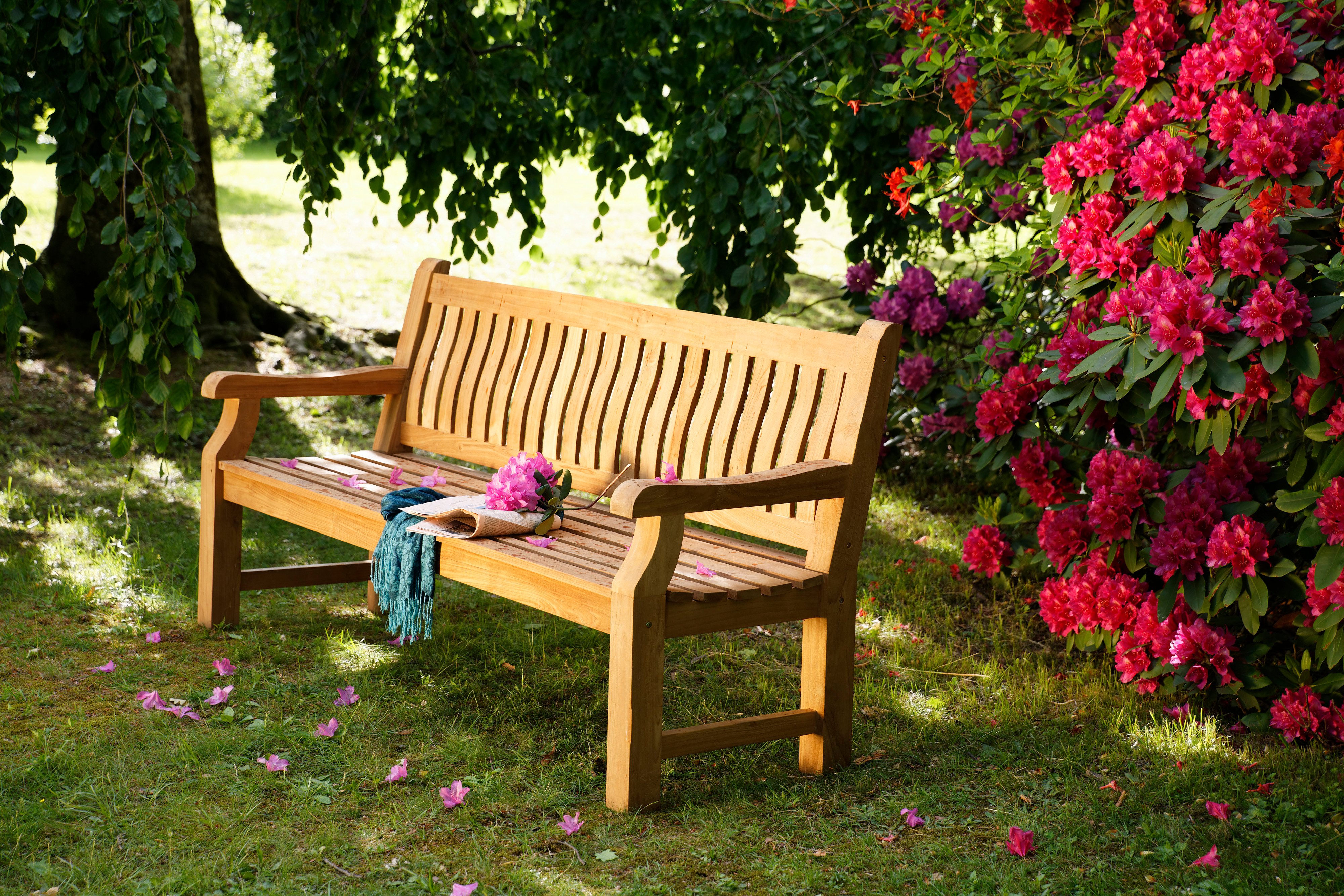 Landhausbank aus recyceltem Teak-Holz im Garten mit blühenden Rhododendren Holzbank aus recyceltem Teak, 2-3 Sitzer, im Garten auf Gras, flankiert von blühendem pinken Rhododendron und einem großen Baum im Hintergrund. Auf der Bank liegt ein rosa Blumenstrauß und ein türkisfarbener Schal.