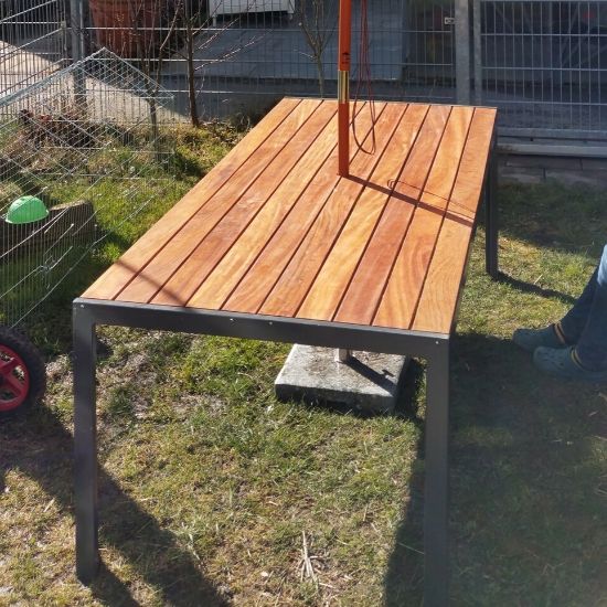Rechteckiger Gartentisch mit hellbraunen Holzplanken und grauem Metallgestell auf Rasen, Schatten eines Pfostens und Kabels auf der Platte, Betonblock darunter, Zaun und Hauswand im Hintergrund, am Bildrand teilweise Schuh und Rad sichtbar