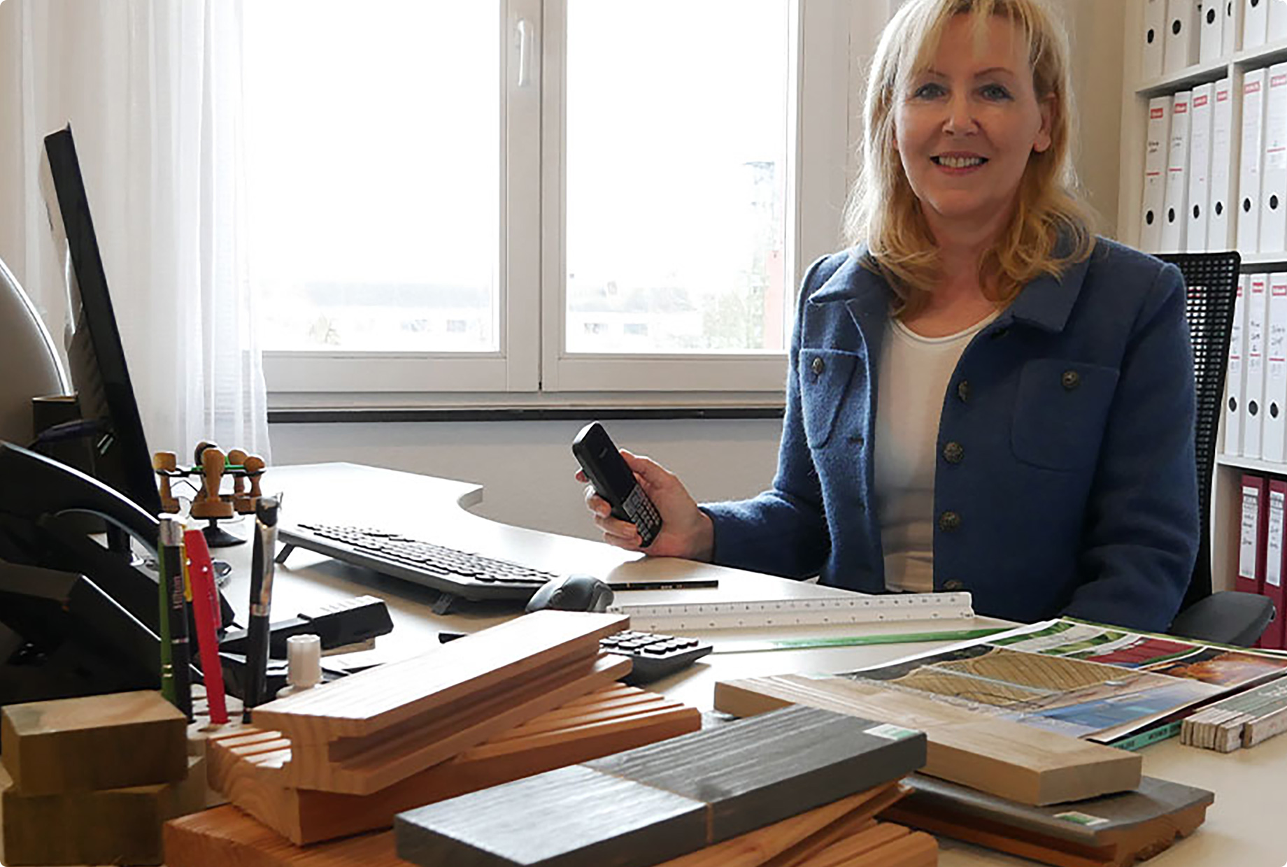 Frau sitzt an einem Büroarbeitsplatz und lächelt, hält einen Telefonhörer; auf dem Schreibtisch liegen Musterhölzer, Prospekte, Tastatur und Stifte, im Hintergrund Fenster und Regale mit Ordnern