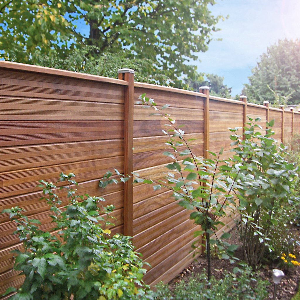 Cumaru Hollandzaun mit profilierten Brettern, ca. 2 x 14 x 200 cm, langlebiger Sichtschutz aus Holz für den Garten Cumaru Hollandzaun mit profilierten Brettern, ca. 2 x 14 x 200 cm, montiert im Garten, gerade verlaufend, dahinter Bäume und blauer Himmel, im Vordergrund grüne Sträucher und Pflanzen