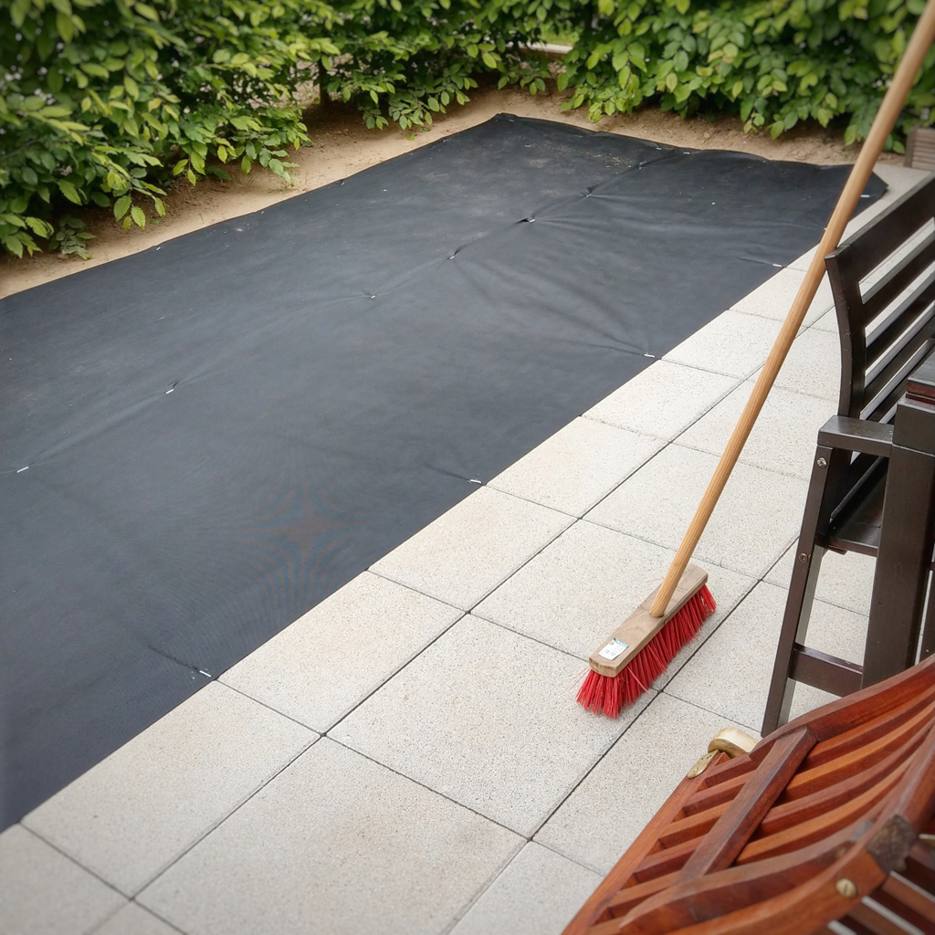 Terrasse mit hellen Betonplatten und großflächiger schwarzer Unkrautfolie entlang einer grünen Hecke; roter Besen mit Holzstiel lehnt auf den Platten neben dunkelbraunen Holzstühlen