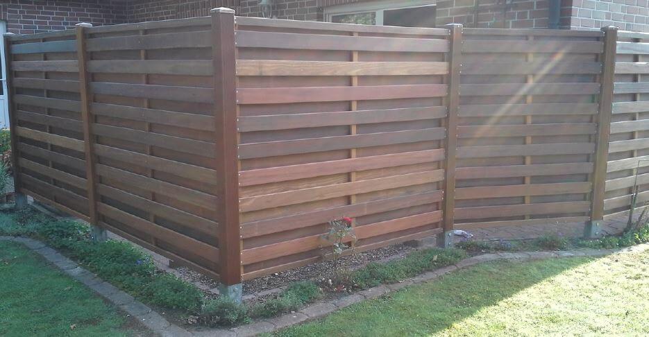 Eckbereich eines horizontalen Lattenzauns aus dunklem Holz vor einer Backsteinwand, kleiner Blumenstreifen mit einer einzelnen Rosenpflanze und niedrigen Bodendeckern, Rasen im Vordergrund und Sonnenreflexe auf dem Zaun