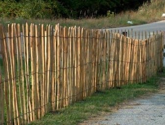 Haselnusszaun Haselnuss Staketenzaun, naturbelassen, 120 cm hoch, aus senkrechten, unregelmäßig geformten Holzlatten und waagerechten Drähten, steht auf einer Wiese am Straßenrand