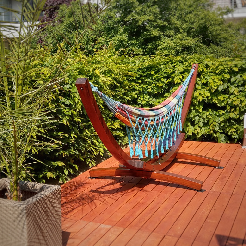 Bunte gestreifte Hängematte mit blauen Netzfransen an gebogenem Holzgestell auf roter Holzterrasse, dichter grüner Heckenkreis im Hintergrund, links ein geflochtener Pflanzkübel mit Palme, sonnige Gartenszenerie