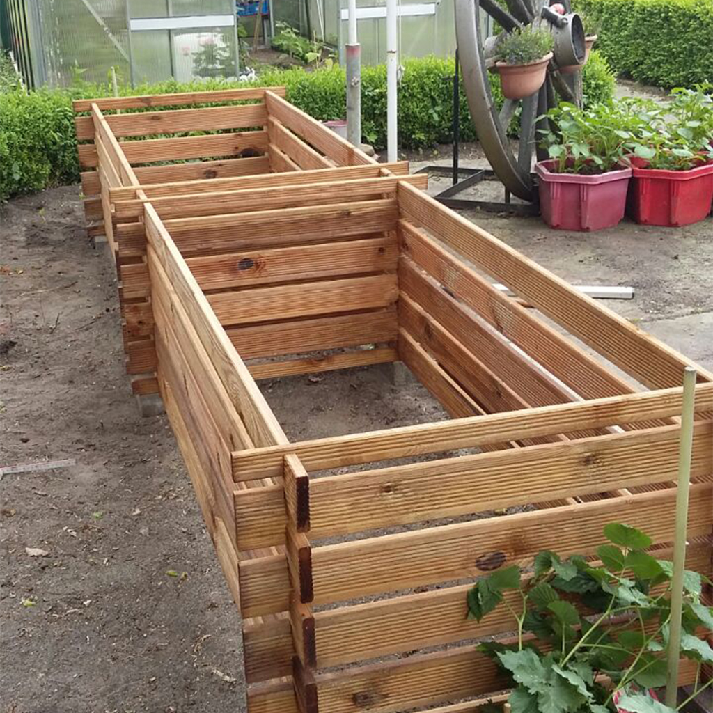 Zwei lange, ineinandergesteckte Hochbeete aus Holzlatten auf Gartenboden, noch nicht bepflanzt; im Hintergrund Gewächshaus, Pflanzkübel und ein dekoratives Wagenrad
