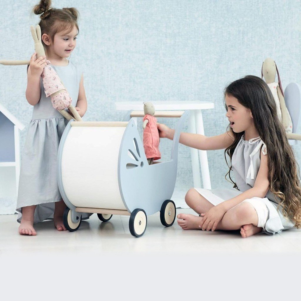 Zwei Kinder spielen mit Puppen und dem Puppenwagen Lilli Zwei Mädchen spielen mit einem hellblauen Puppenwagen Lilli und Stoffpuppen in einem hell eingerichteten Kinderzimmer. Das eine Mädchen steht, das andere sitzt auf dem Boden.
