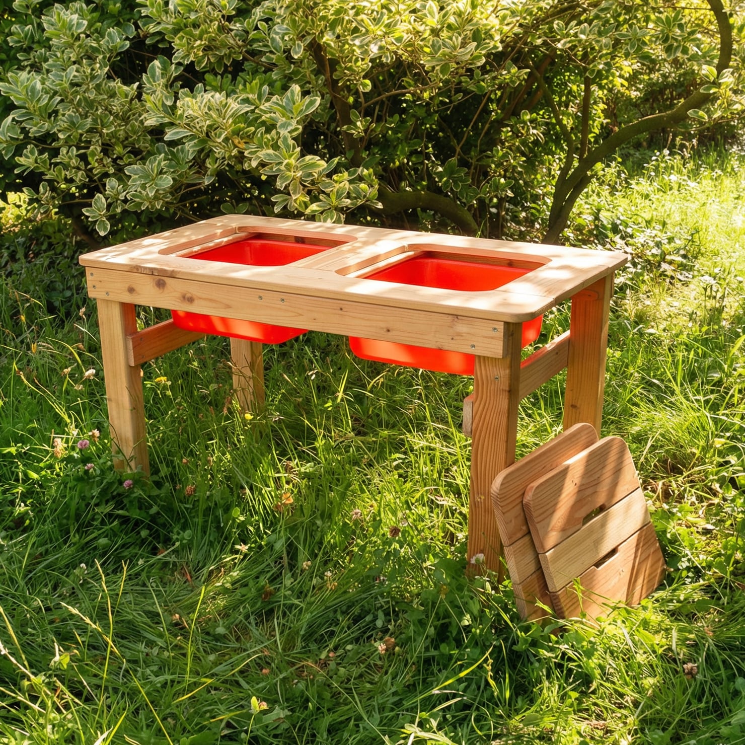 Spieltisch Wassermax Lärche 100 x 50 x 60 cm Spieltisch Wassermax aus Lärche (100×50×60 cm) mit zwei roten Kunststoff-Einsätzen, stabiles Holzgestell und aufgestellte Holzbank, steht im Gras vor einem dichten Strauch