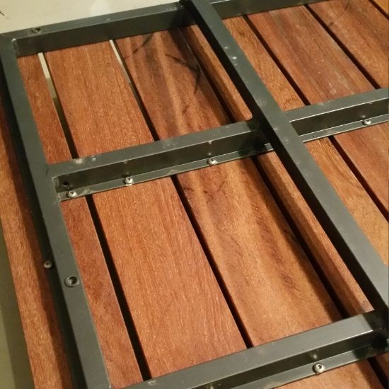 Rechteckiger dunkelgrauer Metallrahmen mit Querverstrebungen und sichtbaren Schrauben liegt auf nebeneinander angeordneten braunen Holzlatten