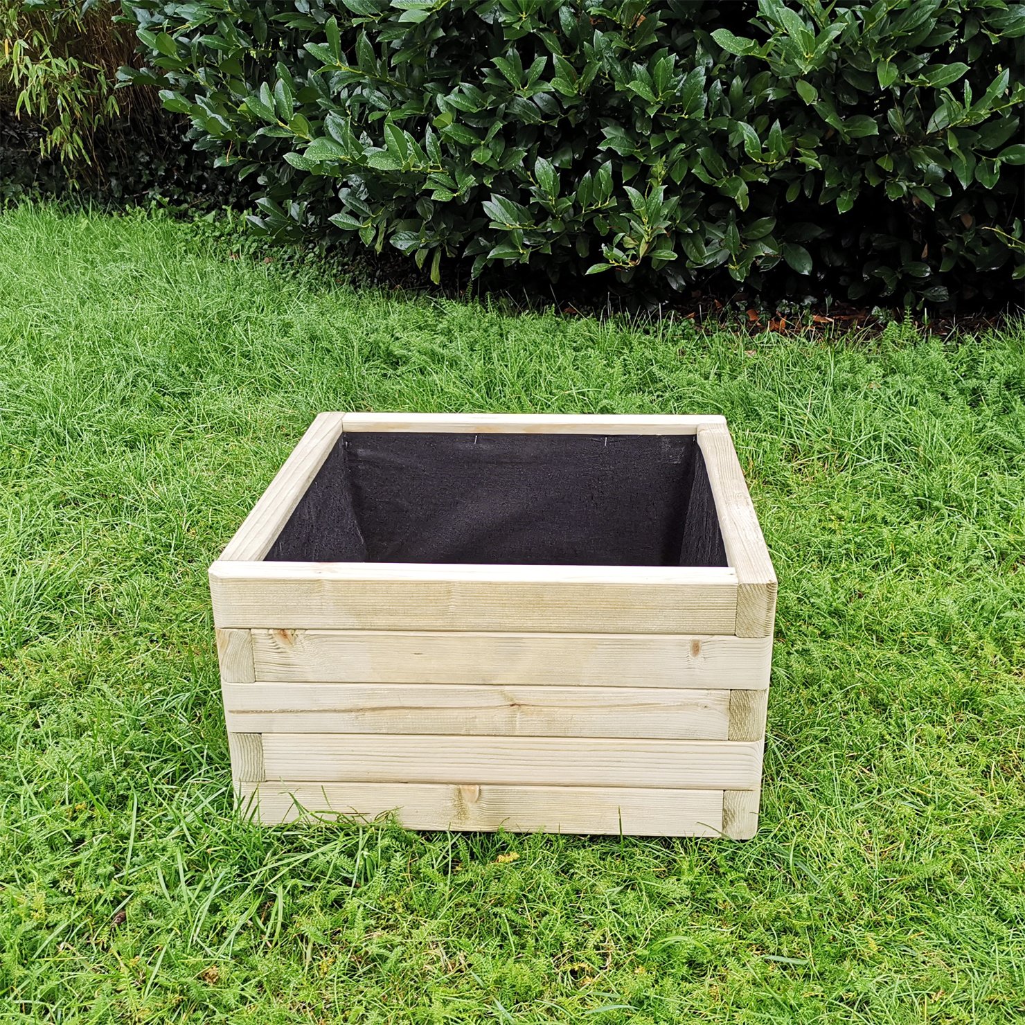 Pflanzkasten Flo aus Fichtenholz mit abgerundeten Ecken für den Garten Quadratischer Pflanzkasten Flo aus Fichtenholz mit abgerundeten Ecken, offene Oberseite mit schwarzer Innenauskleidung, auf grünem Rasen stehend, im Hintergrund dichte grüne Sträucher