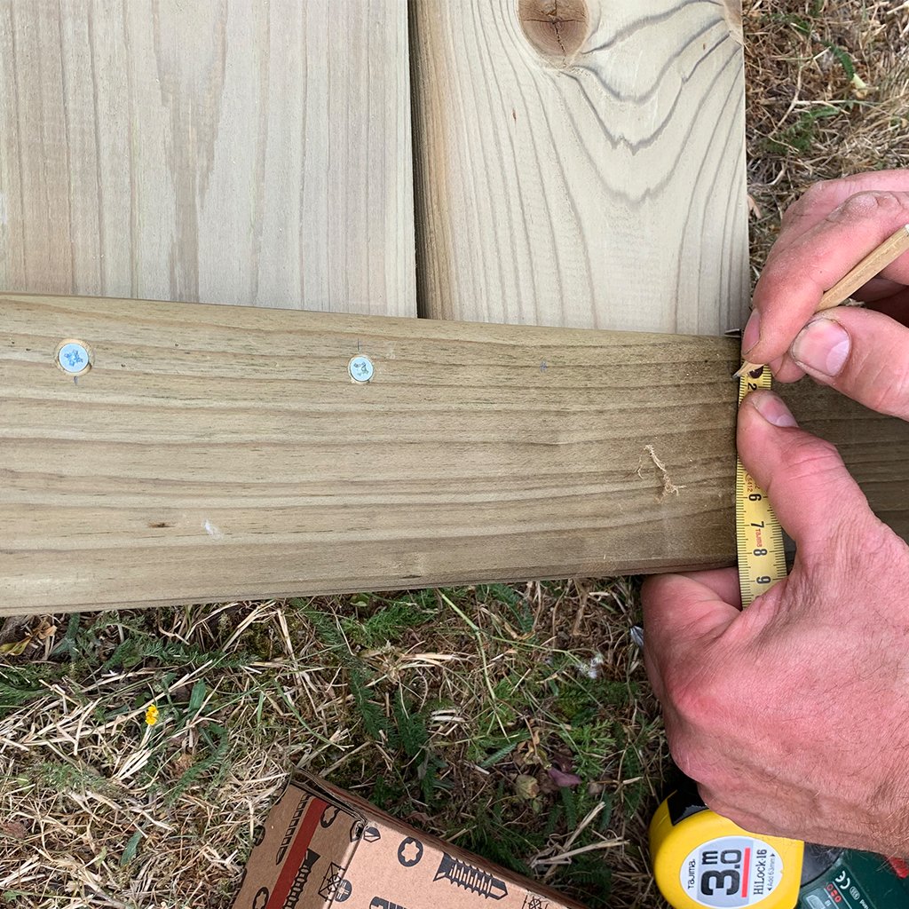Hand misst mit Maßband Holz für den Bau eines individuellen Hochbeets aus Fichte ab Hände messen mit Maßband an einem Hochbeet aus Fichtenholz, verschraubte Holzbretter sichtbar, Rasenuntergrund und Karton mit Schrauben unten im Bild