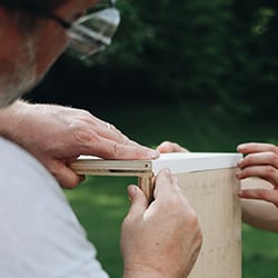 Nahaufnahme: Zwei Personen bei einem DIY-Holzprojekt; eine schleift mit Schleifpapier die Kante eines Holzbretts, die andere hält das Brett fest, unscharfer Gartenhintergrund