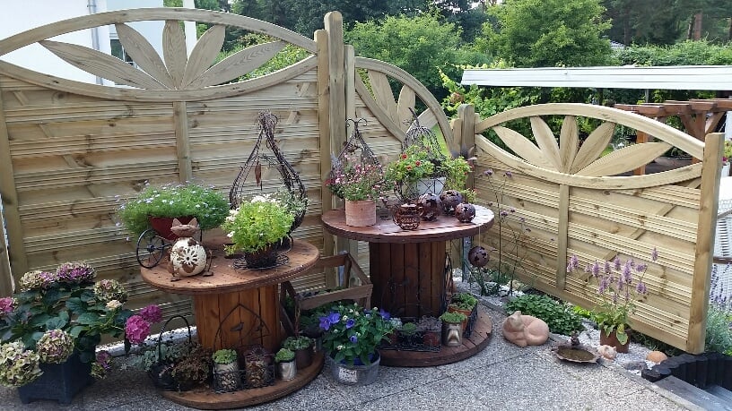 Zaun Verona Ornamentzaun aus kieferngrün imprägniertem Holz als dekorativer Sichtschutz im Garten mit Pflanzen und Gartendekoration Zaun Verona Ornamentzaun aus imprägnierter Kiefer mit dekorativem Sonnenmuster, als Sichtschutz auf Terrasse, davor bepflanzte Kabeltrommeln, verschiedene Blumentöpfe und Gartenaccessoires