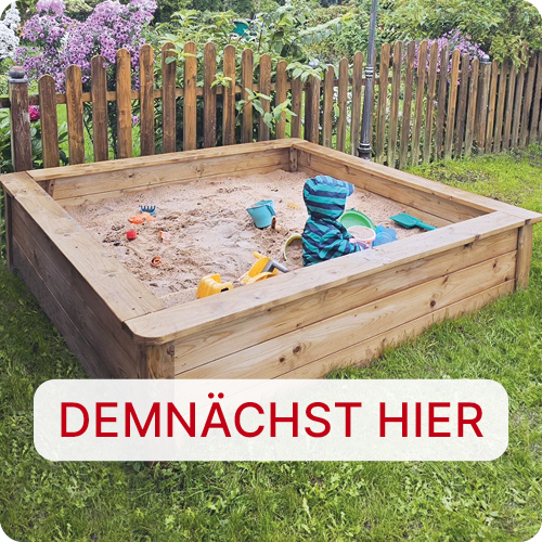 Holz-Sandkasten im Garten mit einem Kind in blau-gestreifter Jacke, Sandspielzeug und Holzzaun vor blühenden Sträuchern; unten im Bild ein weißes Schild mit rotem Schriftzug DEMNÄCHST HIER