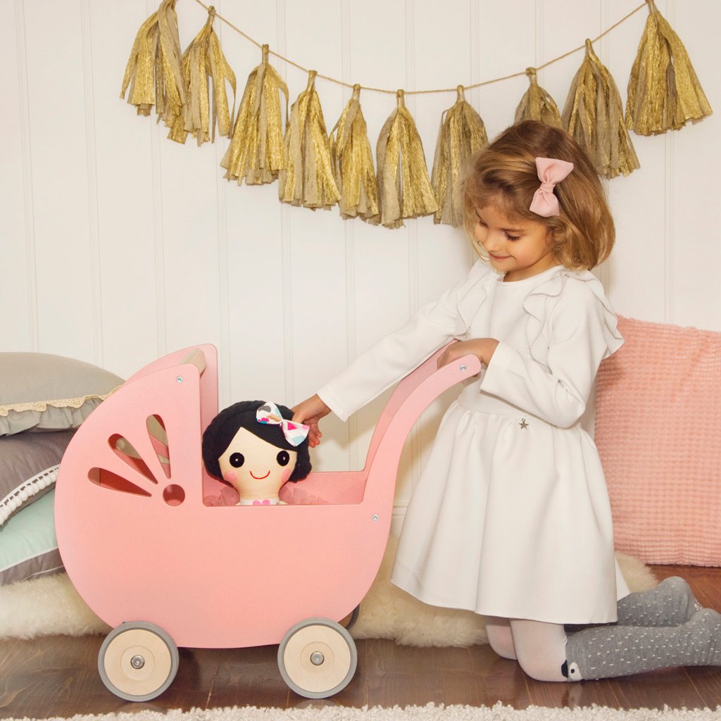 Kleines Mädchen in weißem Kleid spielt mit einem rosa Puppenwagen Lilli aus Holz, in dem eine Stoffpuppe sitzt; im Hintergrund goldene Girlande und gemütliche Kissen.