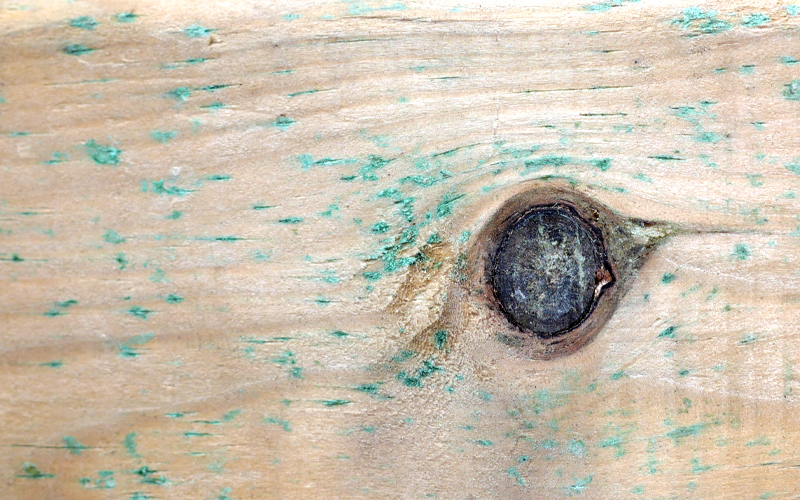 Nahaufnahme eines hellen Holzbretts mit deutlich sichtbarem Astloch rechts der Bildmitte und verstreuten grünen Farbflecken und Abschürfungen auf der Oberfläche